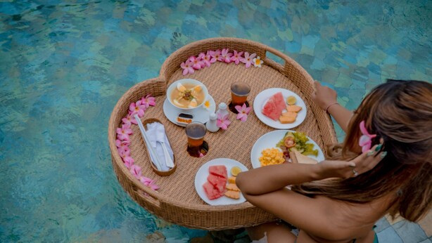 A person sitting by a poolside with a wicker tray containing food and drinks.