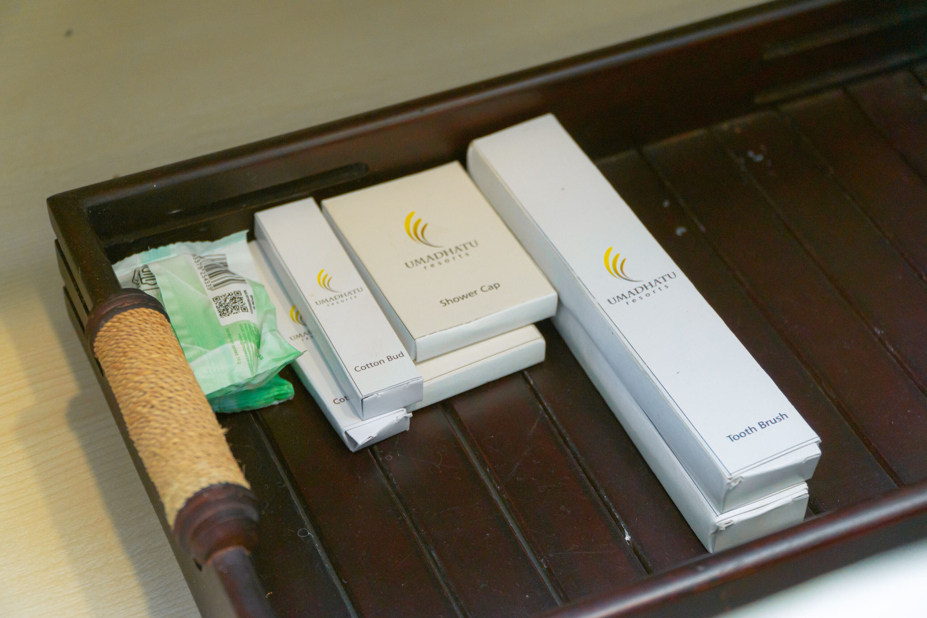 A wooden tray with a wooden-handled brush, a green tissue, and three white boxes labeled "UMADHATU" with "Cotton Bud," "Shower Cap," and "Tooth Brush."