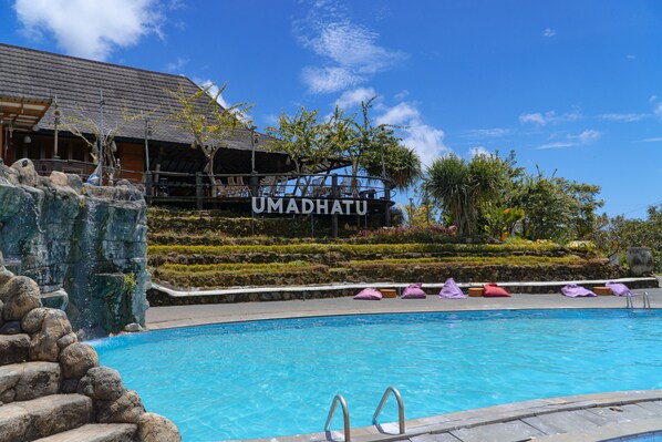 Ein Schwimmbad mit Blick auf ein Gebäude mit der Aufschrift 'UMADHATU' und eine angelegte Gartenanlage.