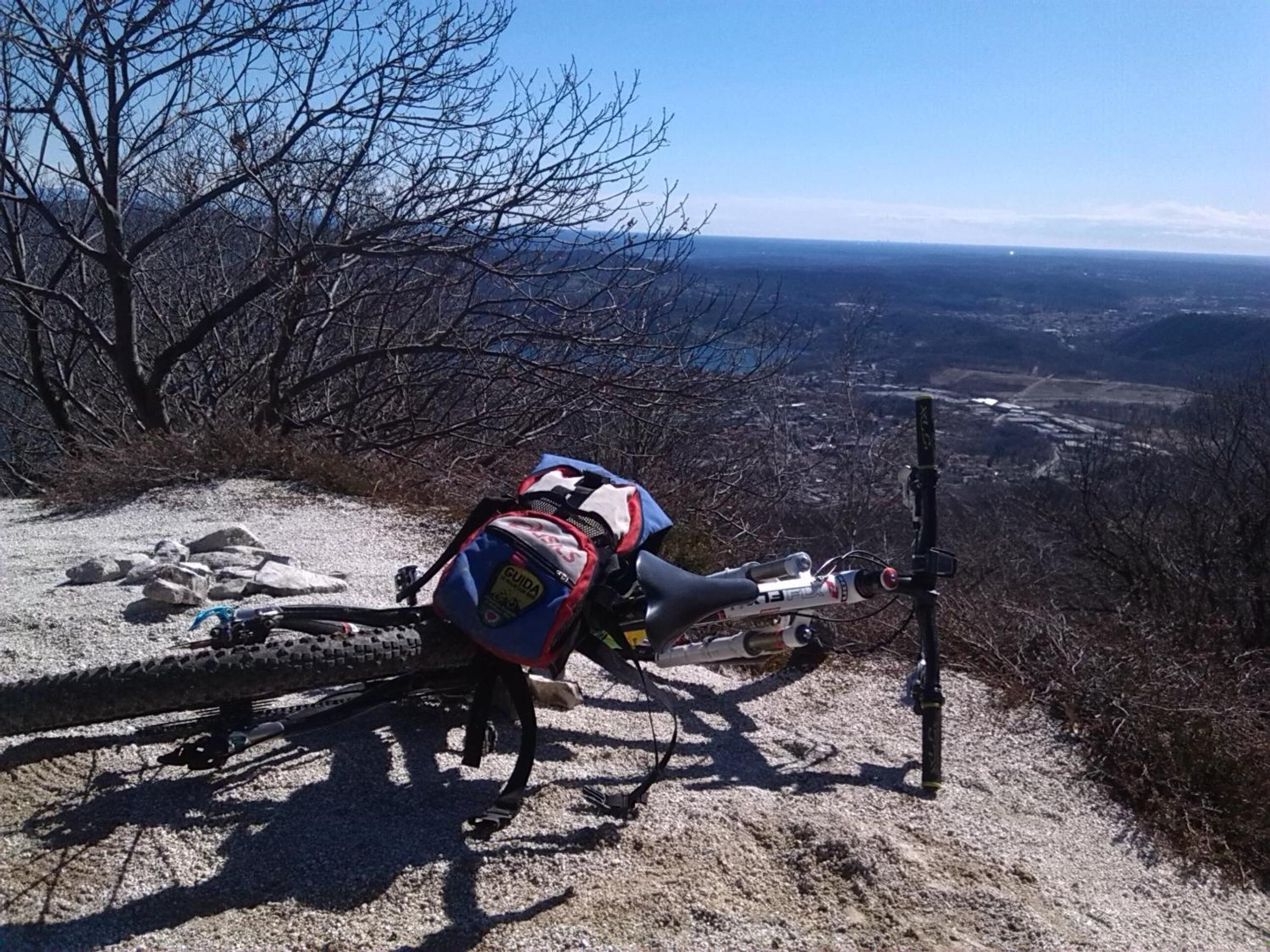 Ein Mountainbike mit blau-rotem Rucksack auf einem Schotterweg, mit Blick auf ein Tal und Berge im Hintergrund.