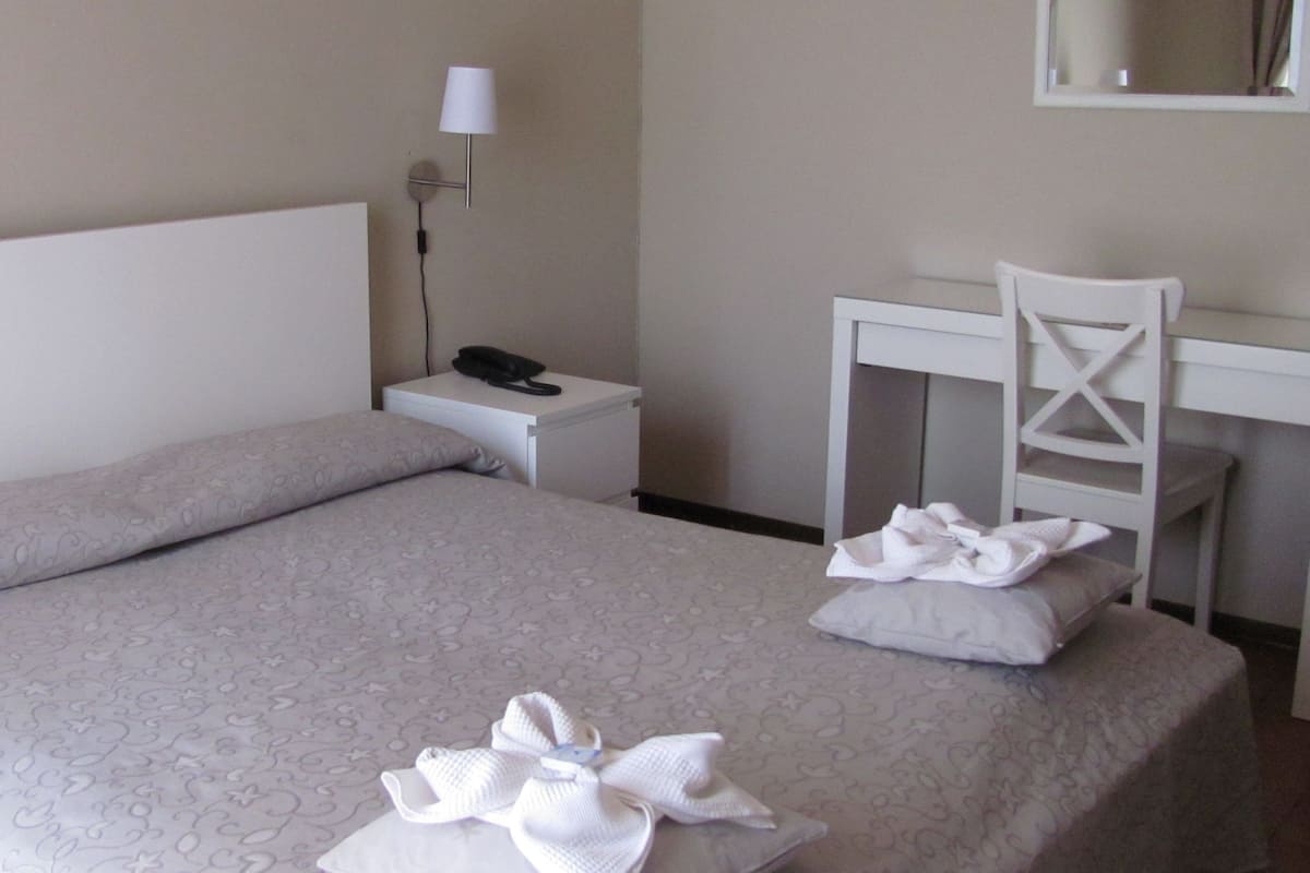A neatly made bed with a patterned bedspread, a bedside telephone, a desk with a chair, and a lamp on the wall.