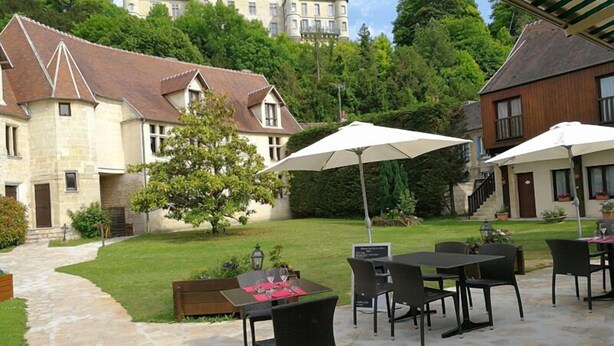 An outdoor patio area with stone flooring, tables, and chairs, surrounded by lush greenery and a large building in the background. - Auberge du jeu darc (Mello)
