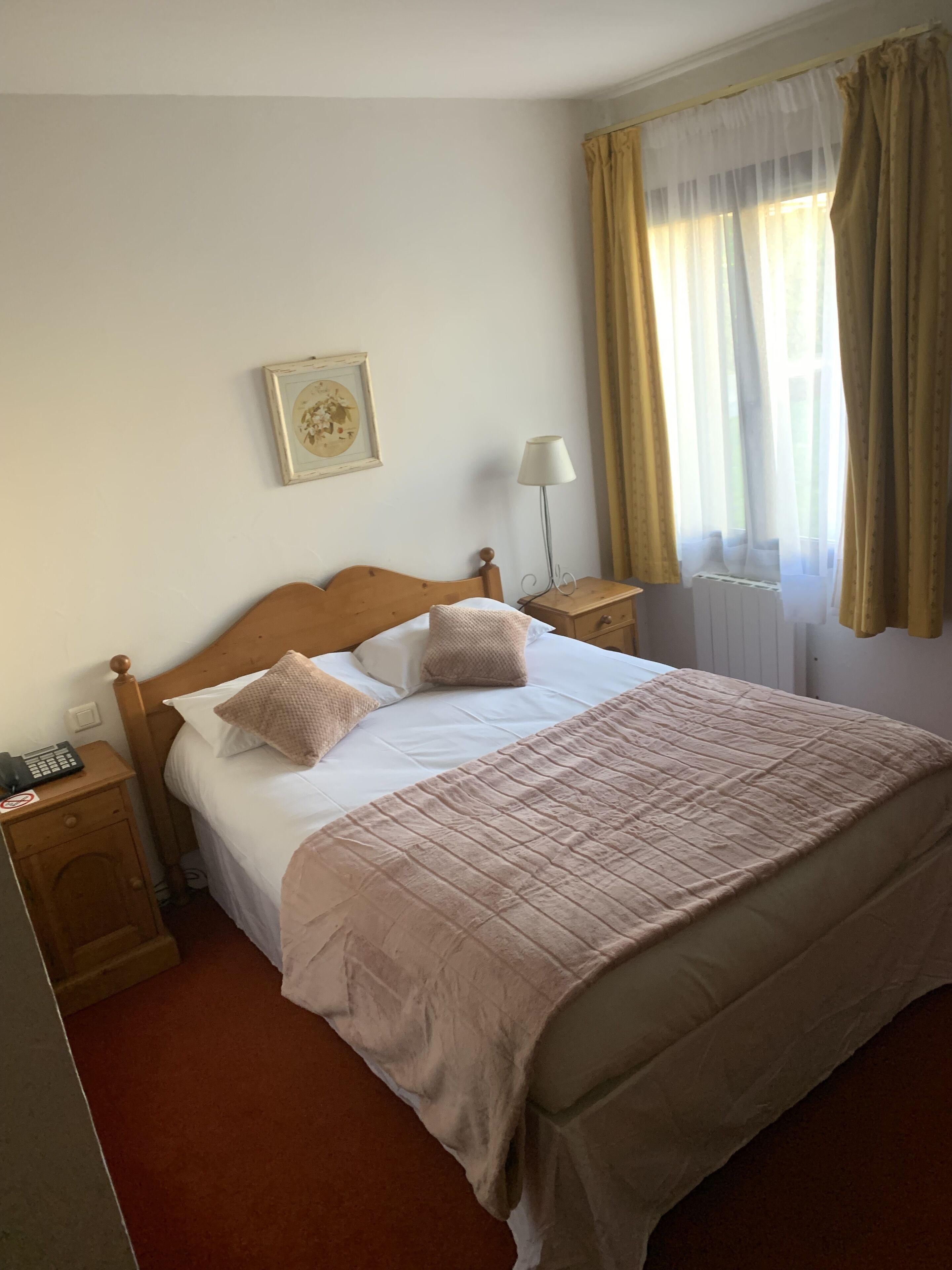 A neatly made bed with a beige comforter and pillows, a bedside lamp, a framed picture on the wall, and a window with curtains.