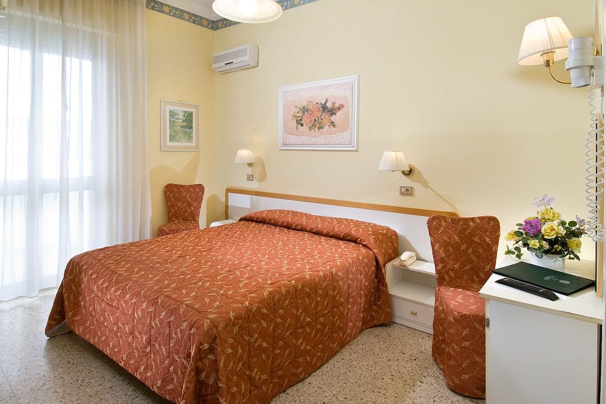 Une chambre d’hôtel avec un lit, une table de chevet, une lampe et un bureau agrémenté d’un vase de fleurs.