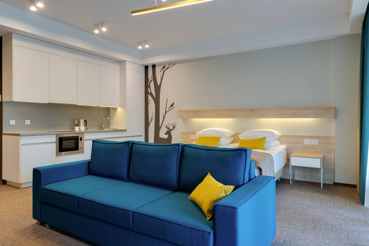 A modern hotel room with a blue sofa, a bed with white and yellow pillows, a small bedside table, and a kitchenette with a microwave.