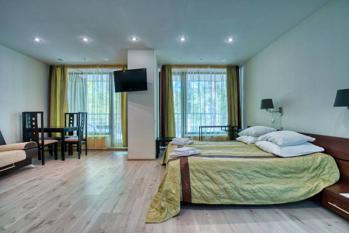 A hotel room with a bed, a TV mounted on the wall, a desk with chairs, and large windows with curtains.