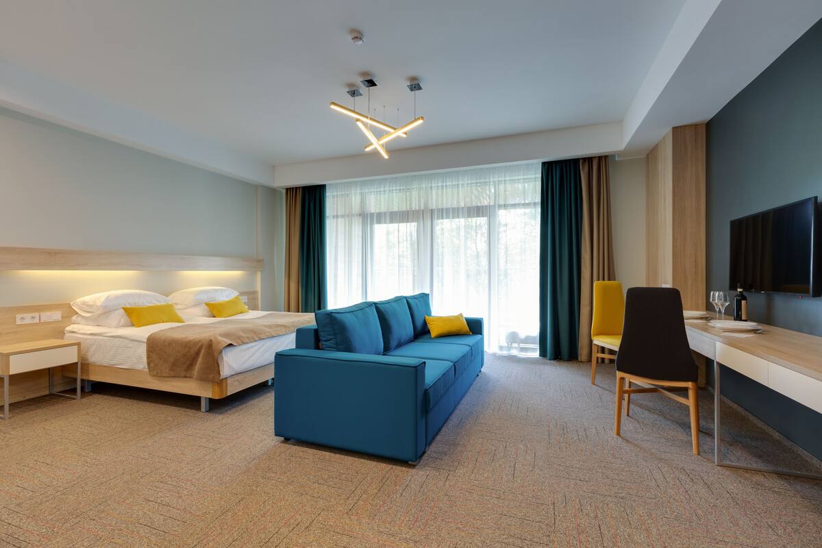 A modern hotel room with a bed, a blue sofa, a desk, and a TV.
