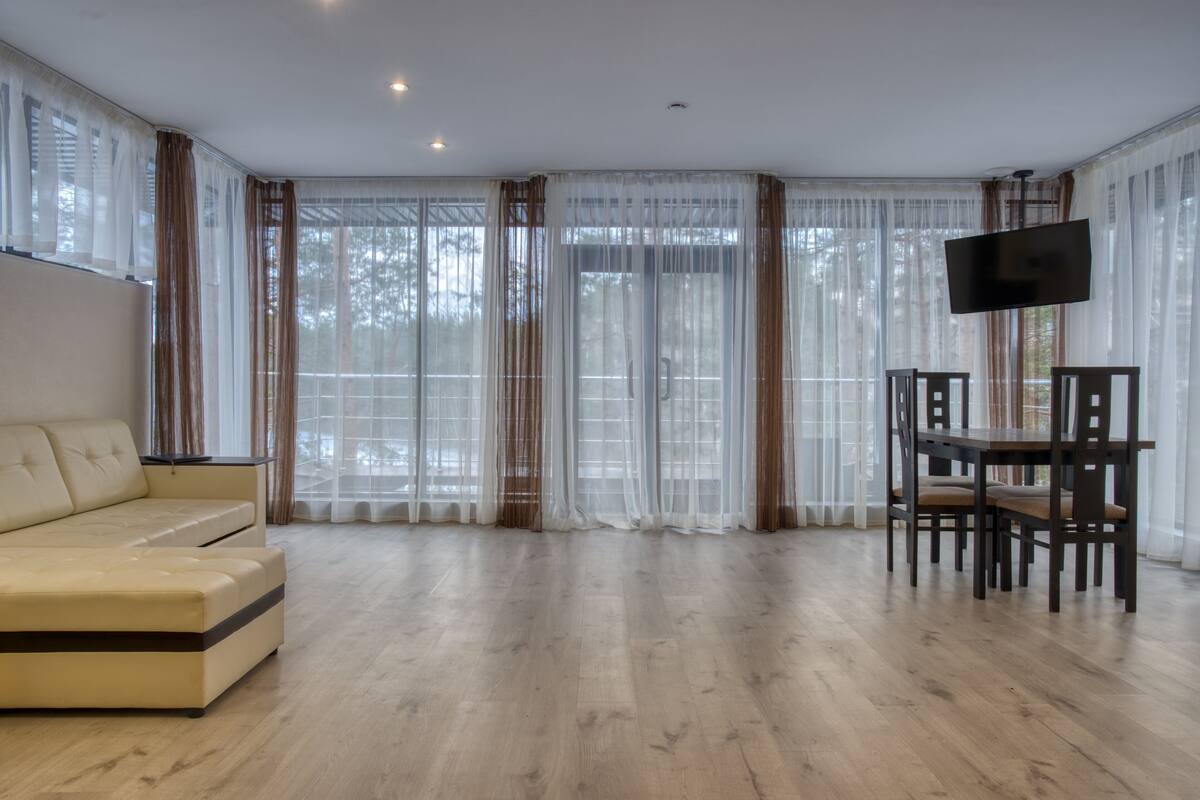 A spacious living room with a beige sofa, a wooden dining table with chairs, and a flat-screen TV mounted on the wall.