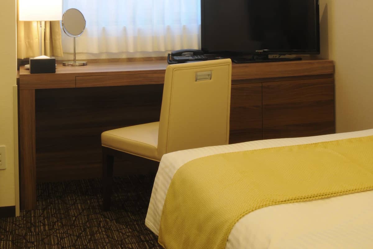 Une chambre d’hôtel avec un lit, un bureau avec une télévision, une chaise, une lampe et une fenêtre avec des rideaux.