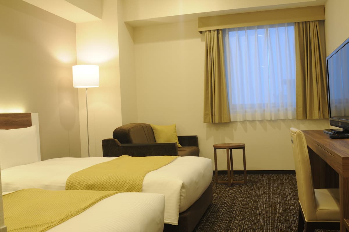 Une chambre d’hôtel avec deux lits, un bureau, une chaise, une lampe et une fenêtre avec des rideaux.