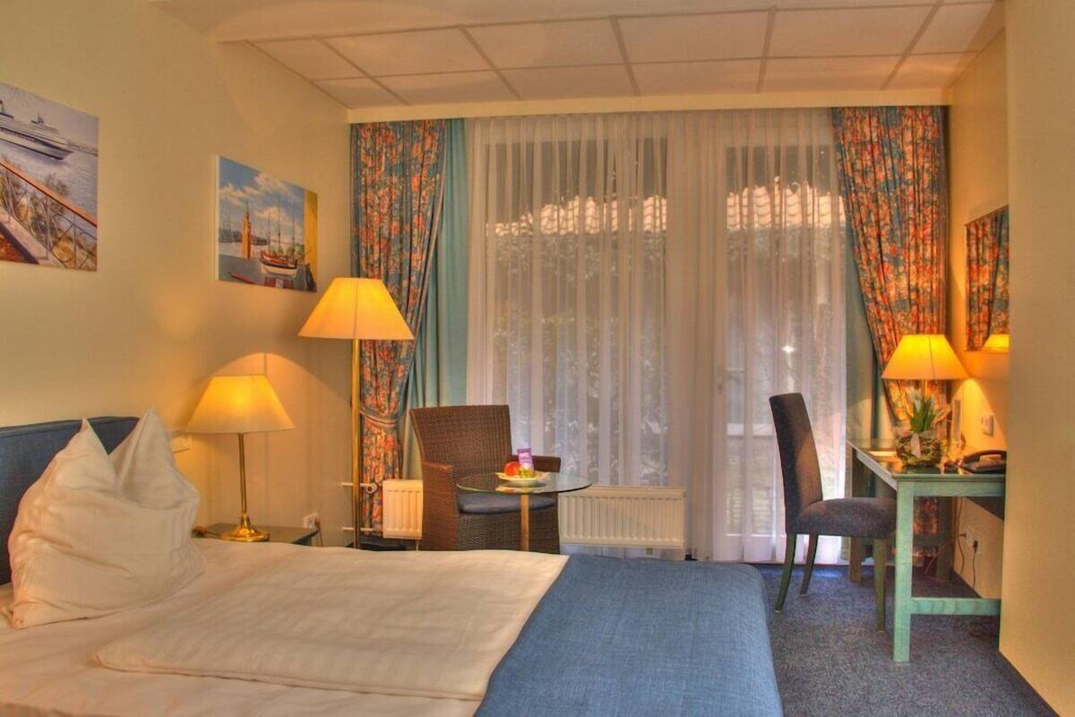 A hotel room with a bed, two chairs, a small table, a desk, and a window with curtains.