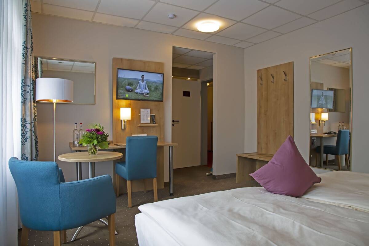 A hotel room with a bed, two chairs, a small table with a lamp, a TV on the wall, and a wooden door.