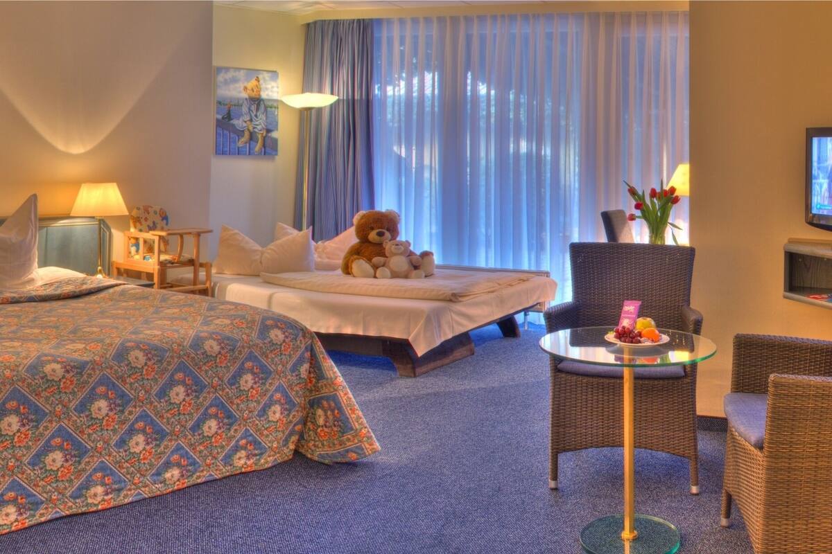 A hotel room with a large bed, two bedside tables with lamps, a small table with fruit, a sofa, a television, and a window with curtains.