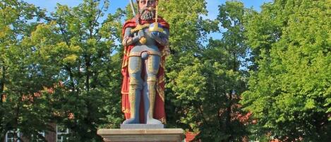 Una estatua de una figura con armadura, una corona y un cetro, sobre un pedestal de ladrillo con un escudo de armas.