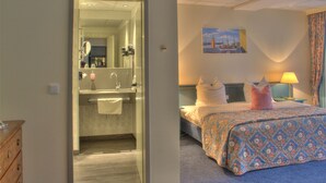 Una habitación de hotel con baño, una cama con cubrecama floral, una mesita de noche con lámpara y un cuadro en la pared.