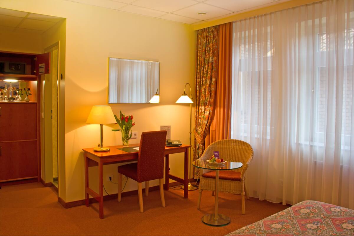 A hotel room with a bed, a desk with a chair, a small table with a chair, a lamp, and a window with curtains.