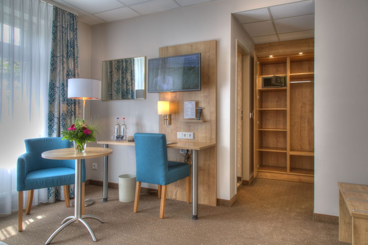 A hotel room with a desk, two chairs, a TV, a lamp, a small table with a vase of flowers, and a wooden wardrobe.
