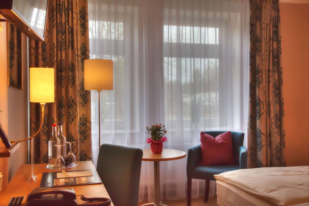 A hotel room with a bed, a desk with a lamp, a chair, a small table with a vase, and curtains.
