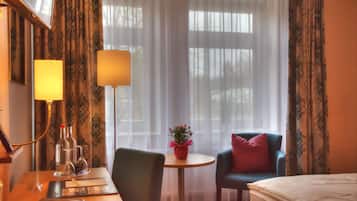 Ein Hotelzimmer mit Bett, Schreibtisch mit Lampe, Stuhl, kleinem Tisch mit Vase und Vorhängen.