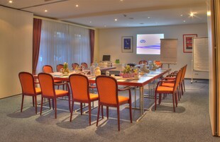 Ein Konferenzraum mit einem langen Tisch, mehreren Stühlen, einer Projektionsleinwand und einer Tafel.