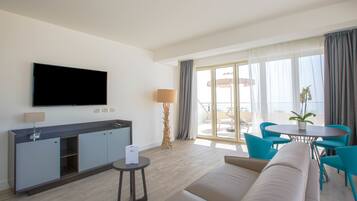 A modern hotel room with a flat-screen TV, a sofa, a dining table with chairs, and a balcony with a view.