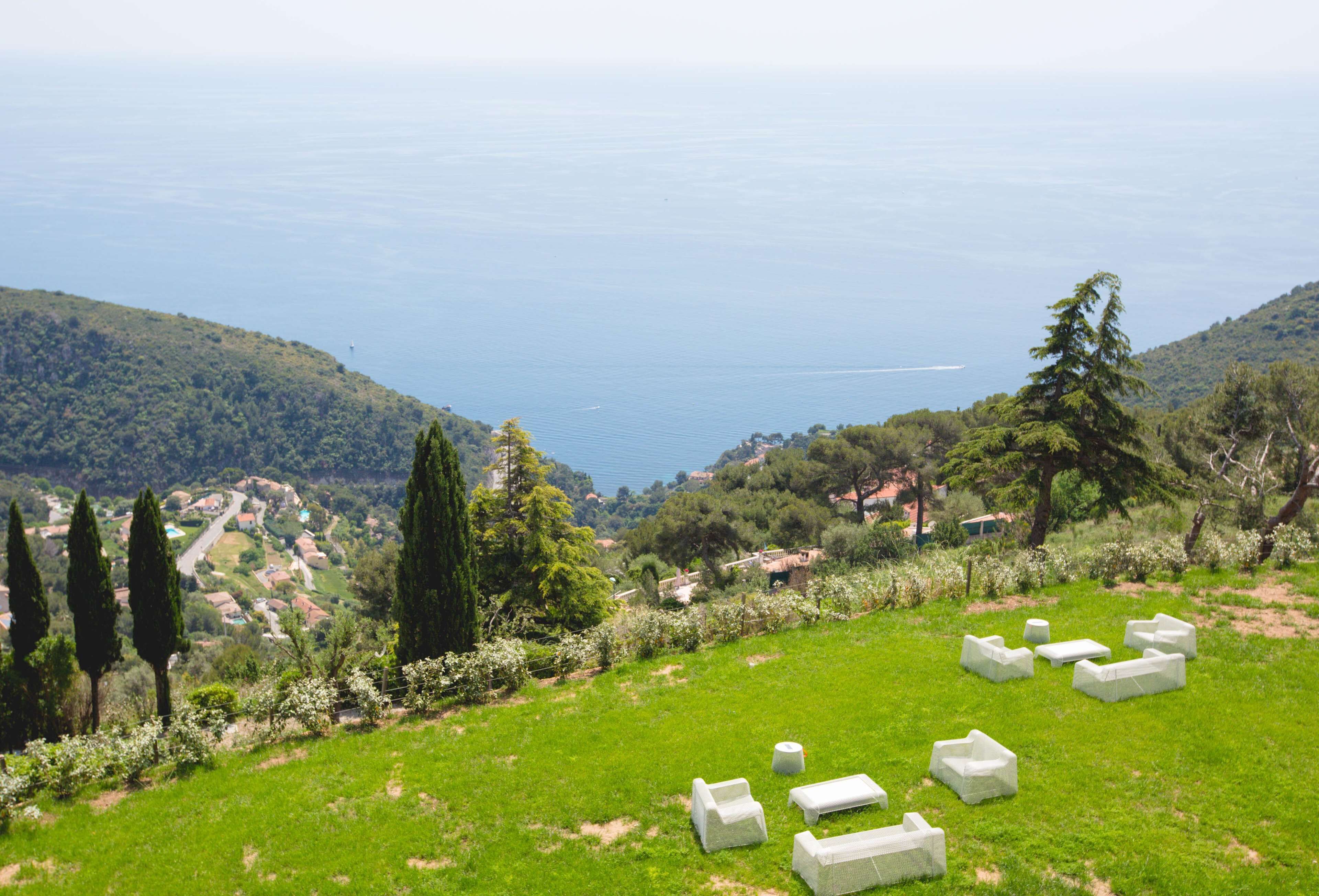 A grassy area with white outdoor furniture arranged in a semi-circle, overlooking a coastal landscape with hills and a body of water.