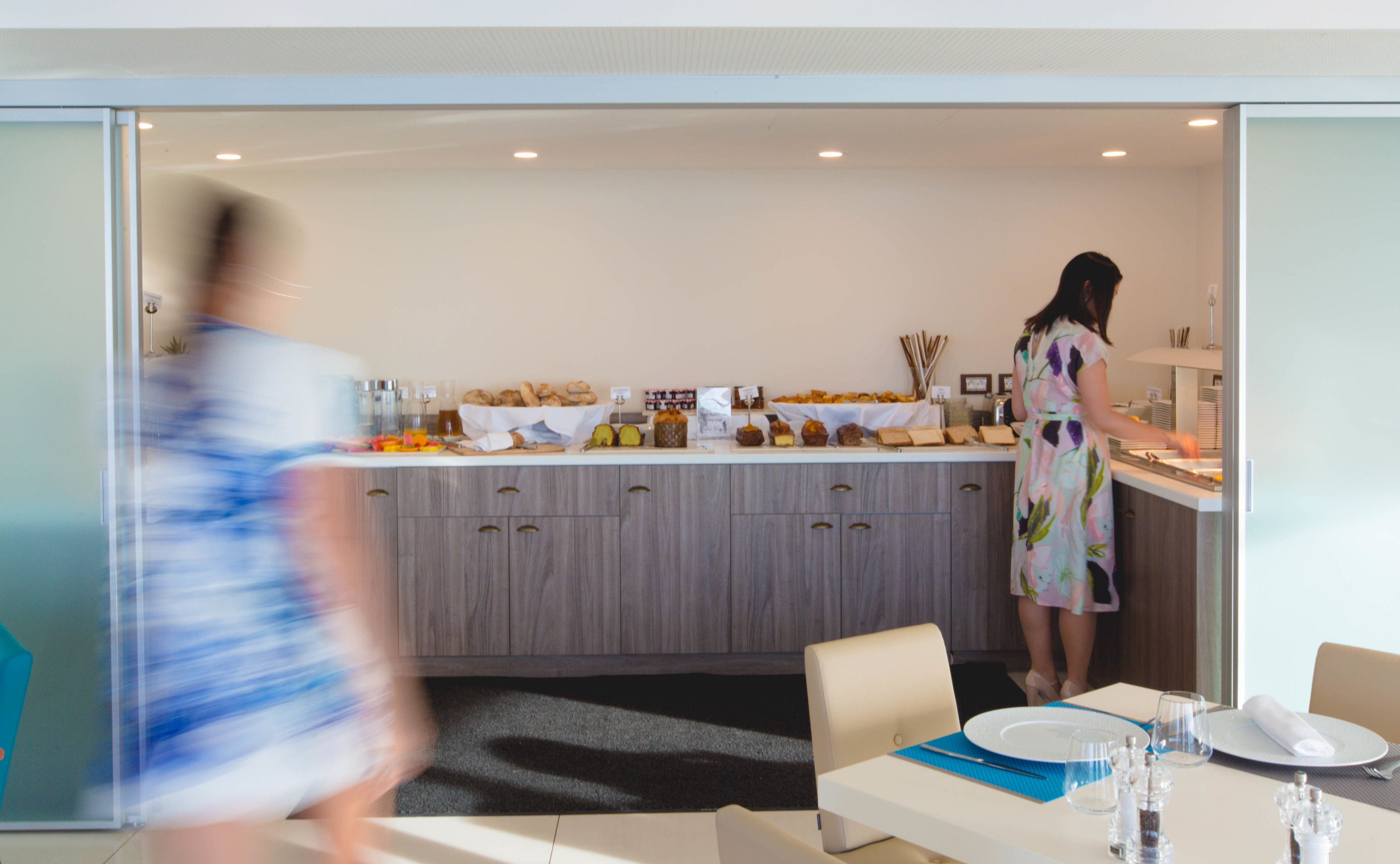 A modern kitchen with a buffet setup, a dining area with a table set for two, and a woman preparing food.