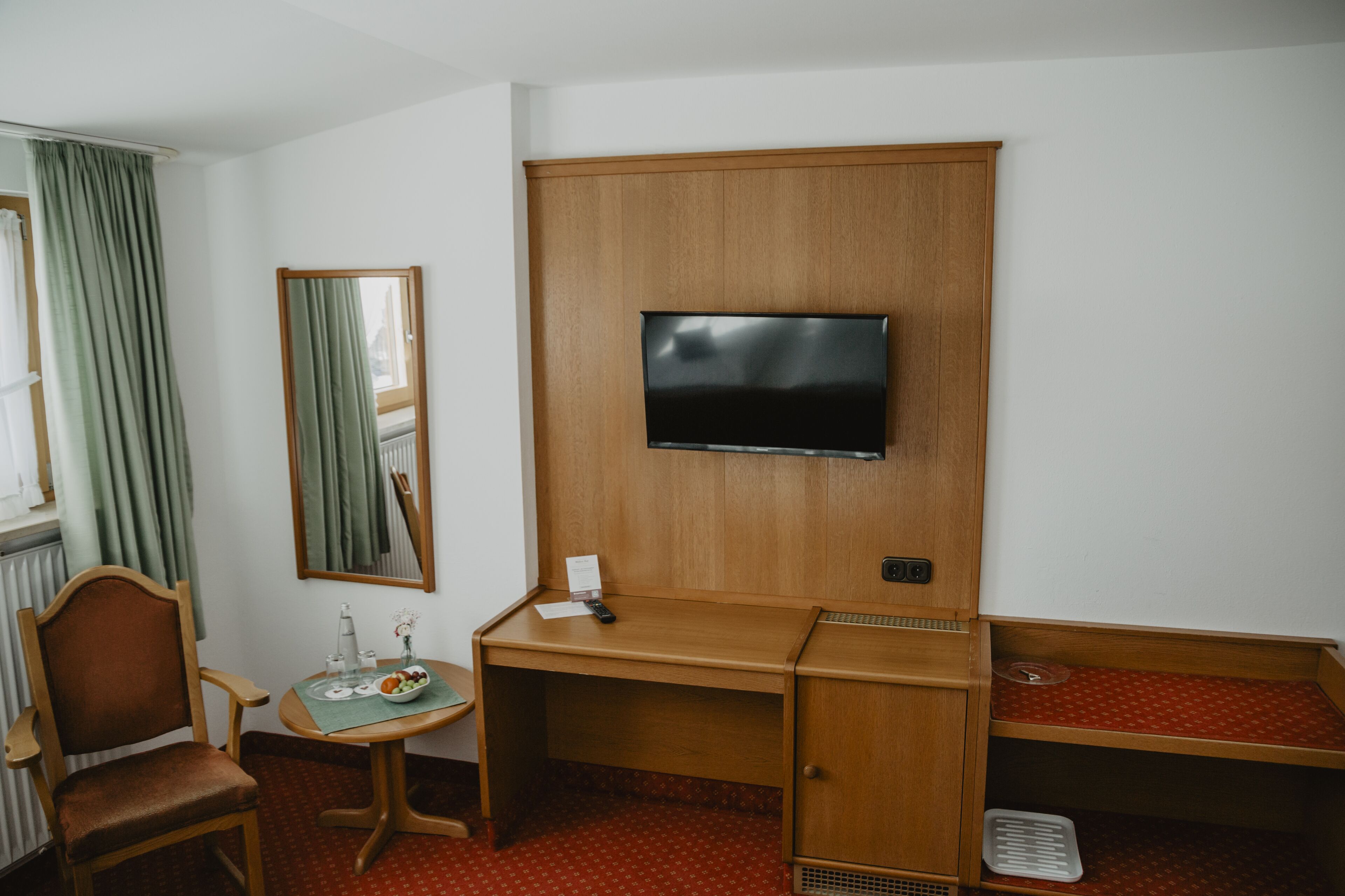 Una habitación de hotel con un escritorio de madera, un televisor montado en la pared, una silla, una mesita con un florero y una fuente de fruta, y un radiador.