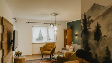 A living room with a sofa, armchair, and a coffee table. There is a window with curtains, a wall-mounted shelf, and a painting.