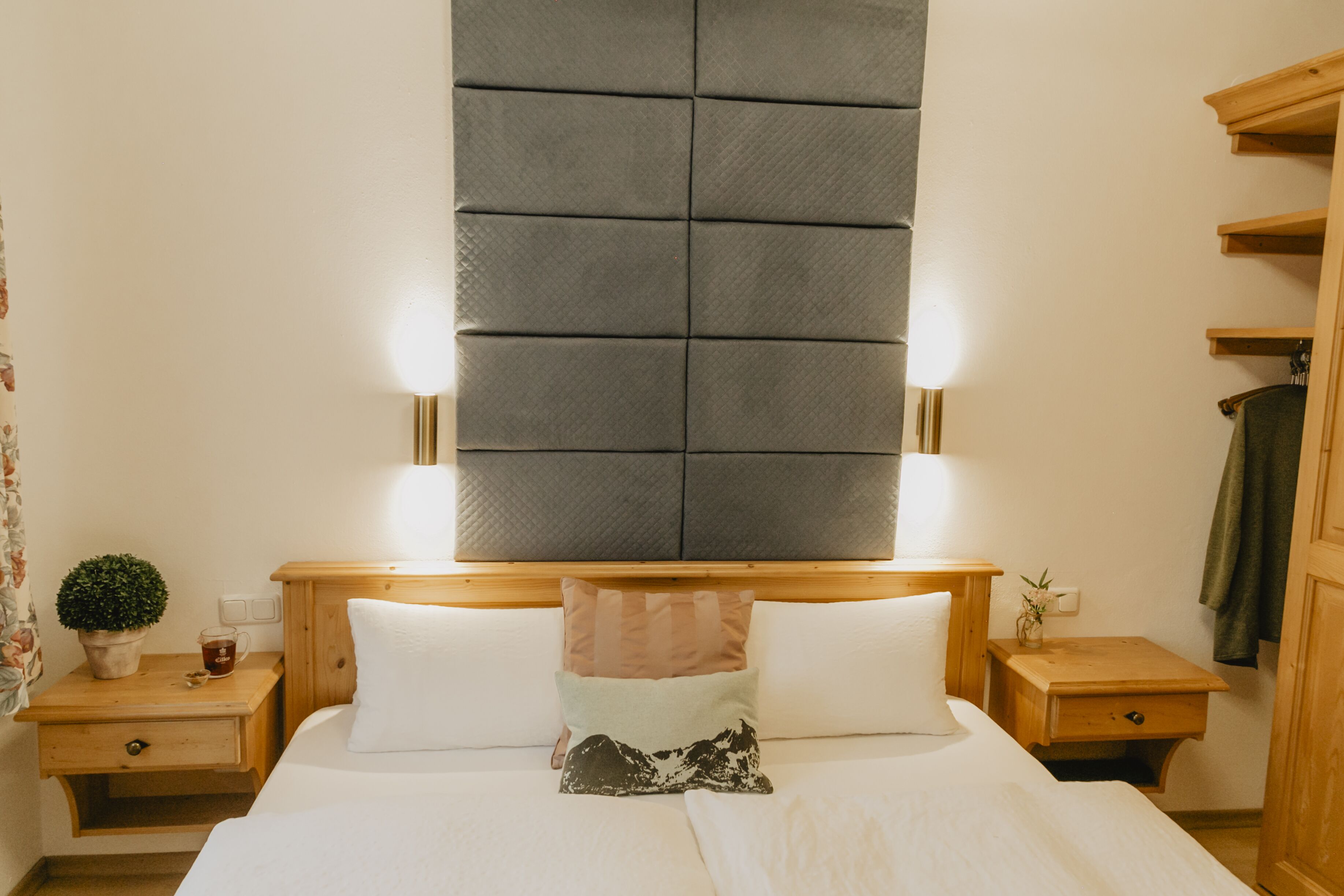 A neatly arranged hotel room with a large bed, two bedside tables, a nightstand, a wooden headboard with built-in lighting, and a small potted plant.