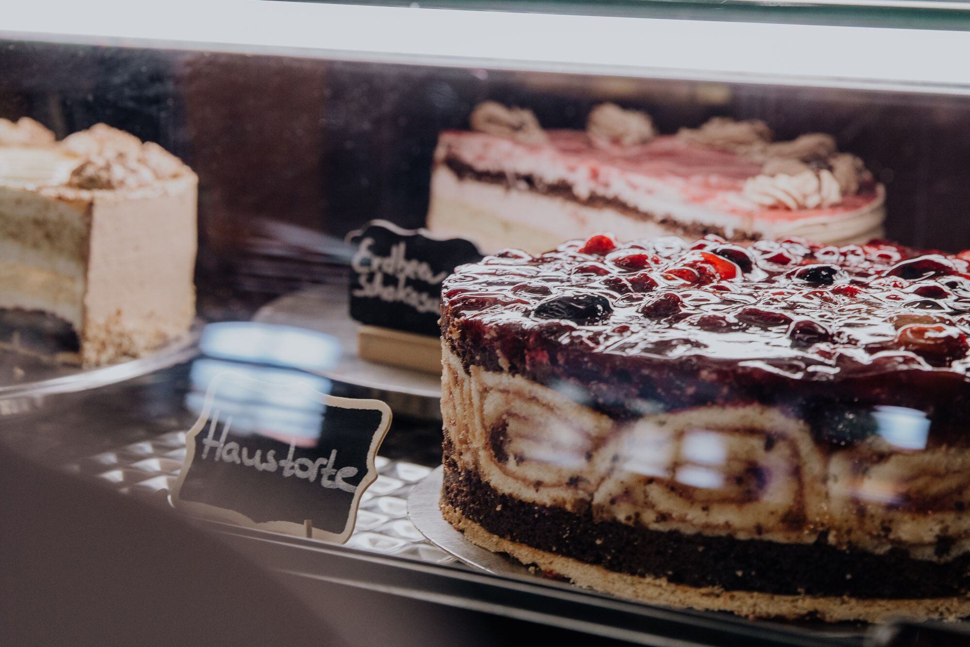Una vitrina con varios pasteles, incluyendo un pastel de chocolate con cobertura de cereza y un pastel con glaseado blanco y decoración de cerezas.