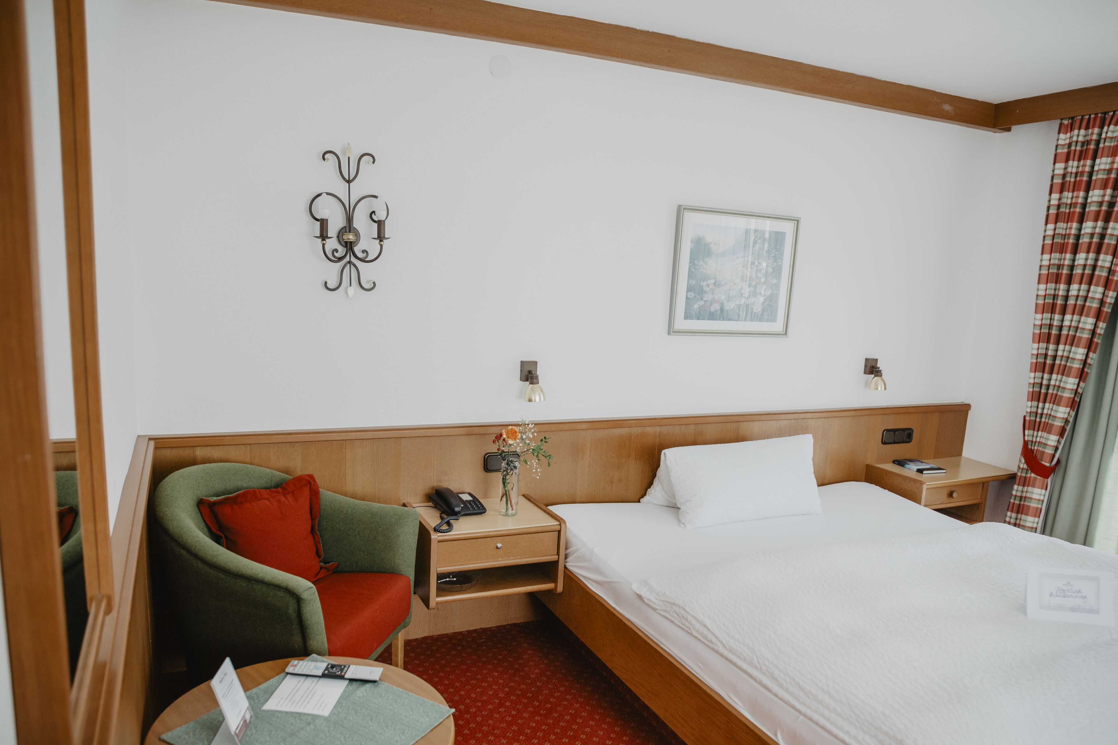 Ein Hotelzimmer mit einem Bett, einem grünen Sessel, einem Holznachttisch, einem kleinen Tisch, einem an der Wand montierten Telefon und einem gerahmten Bild.