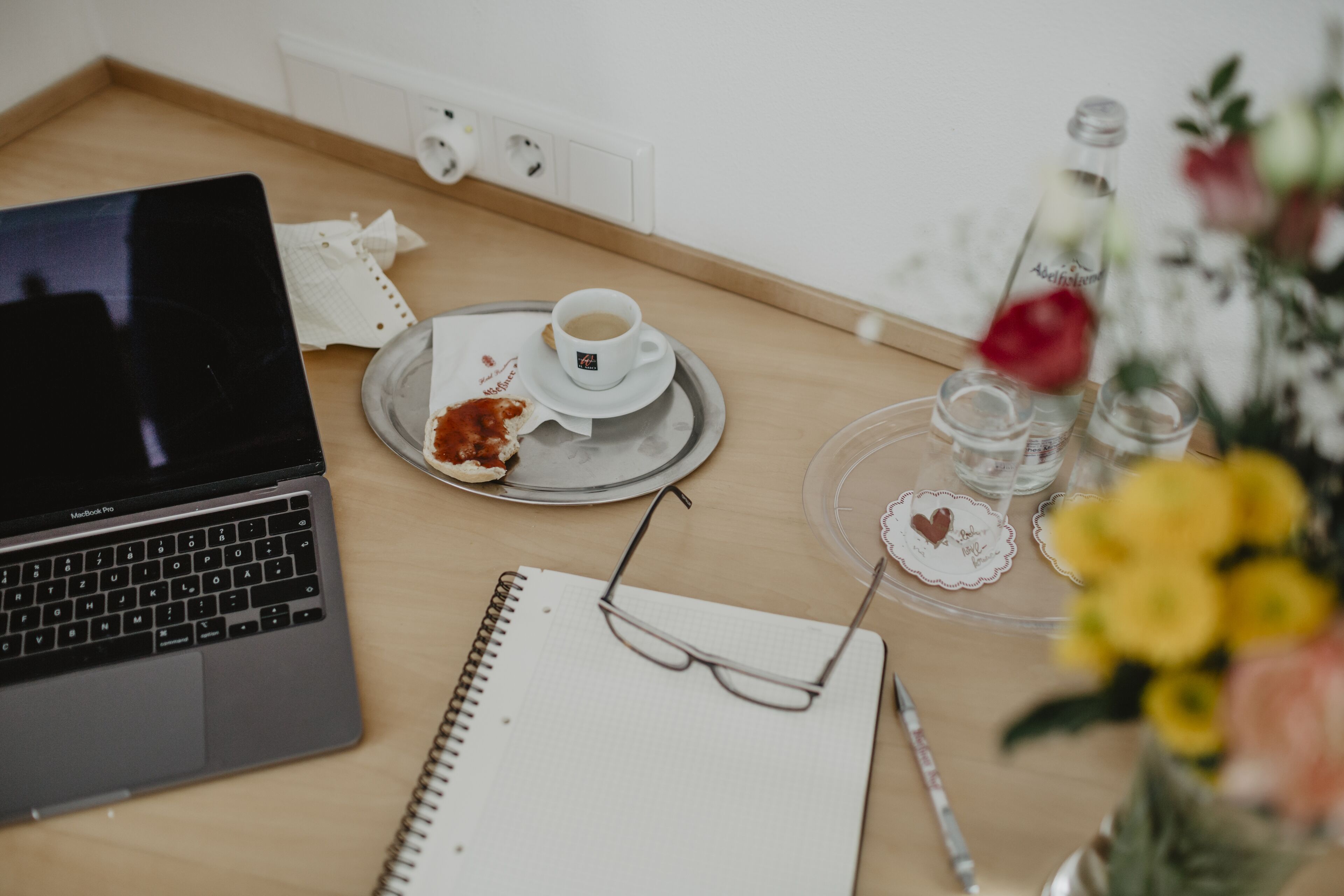 A laptop, a notebook, a pair of glasses, a cup of coffee, a plate with food remnants, and a vase with flowers on a wooden table.