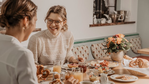 Una mesa de comedor con diversos alimentos para el desayuno, un jarrón de flores y una estantería al fondo.
