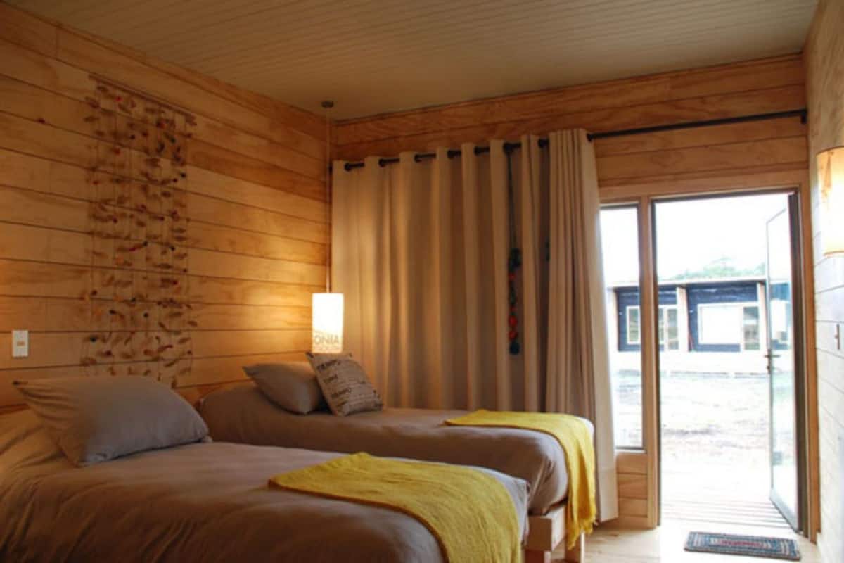 A wooden cabin room with two beds, a wall-mounted light, and a sliding glass door leading to an outdoor area.