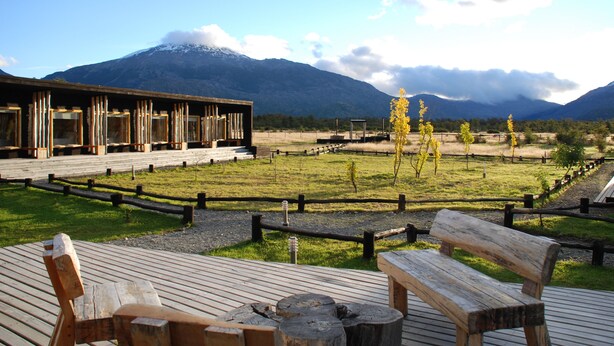 Una terraza de madera con bancos, un área cercada y un edificio con ventanales amplios.