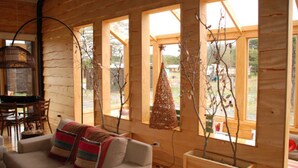 Interior acogedor de una cabaña con sofá, mesa de comedor y sillas. Amplias ventanas permiten luz natural y vistas del exterior.