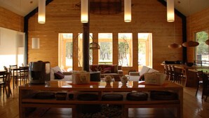 Una habitación amplia con techo alto, paredes de madera y ventanas grandes. Se aprecia una mesa central con diversos objetos encima y una chimenea en el centro.