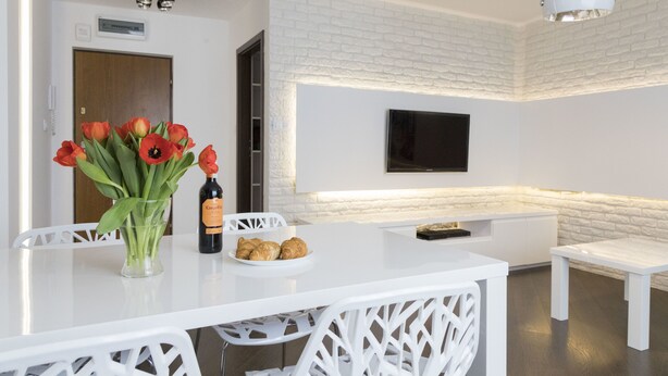 Cozinha moderna com armários brancos, bancada branca, um vaso com flores vermelhas, uma garrafa de vinho e um prato com croissants.