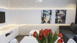 Uma sala de estar moderna com uma parede de tijolos brancos, uma televisão de tela plana montada acima de um console branco, um sofá cinza e uma pequena mesa branca com um vaso de tulipas vermelhas.