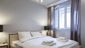 Cama bem arrumada com lençóis brancos e duas toalhas dobradas, mesa de cabeceira com abajur e janela com cortinas.