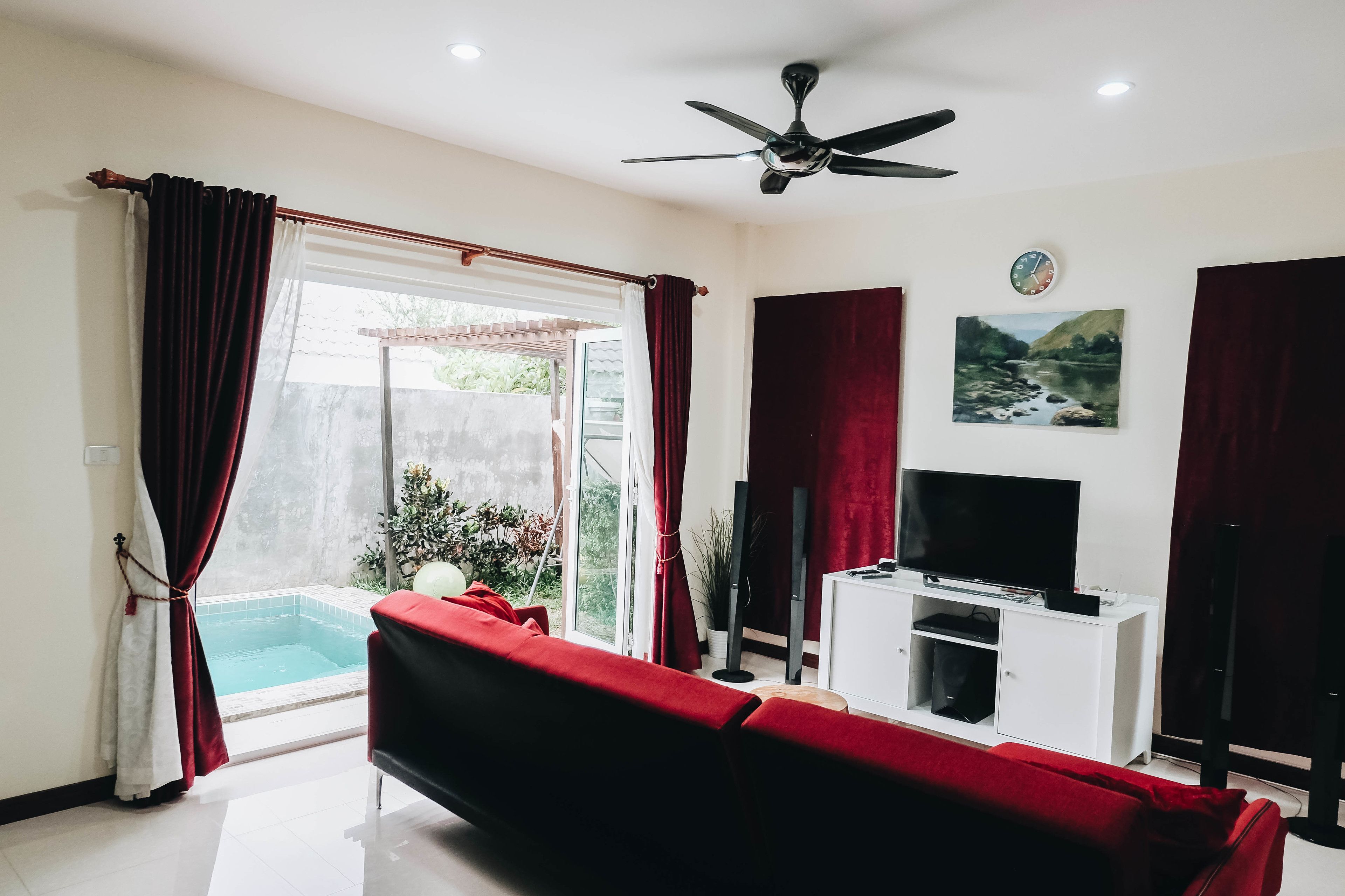 A living room with a red sofa, a flat-screen TV on a white cabinet, a ceiling fan, and a small pool visible through the glass door.