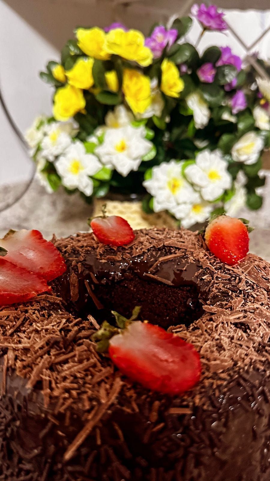 A chocolate cake with strawberries and chocolate shavings on top, placed in front of a bouquet of yellow and white flowers.