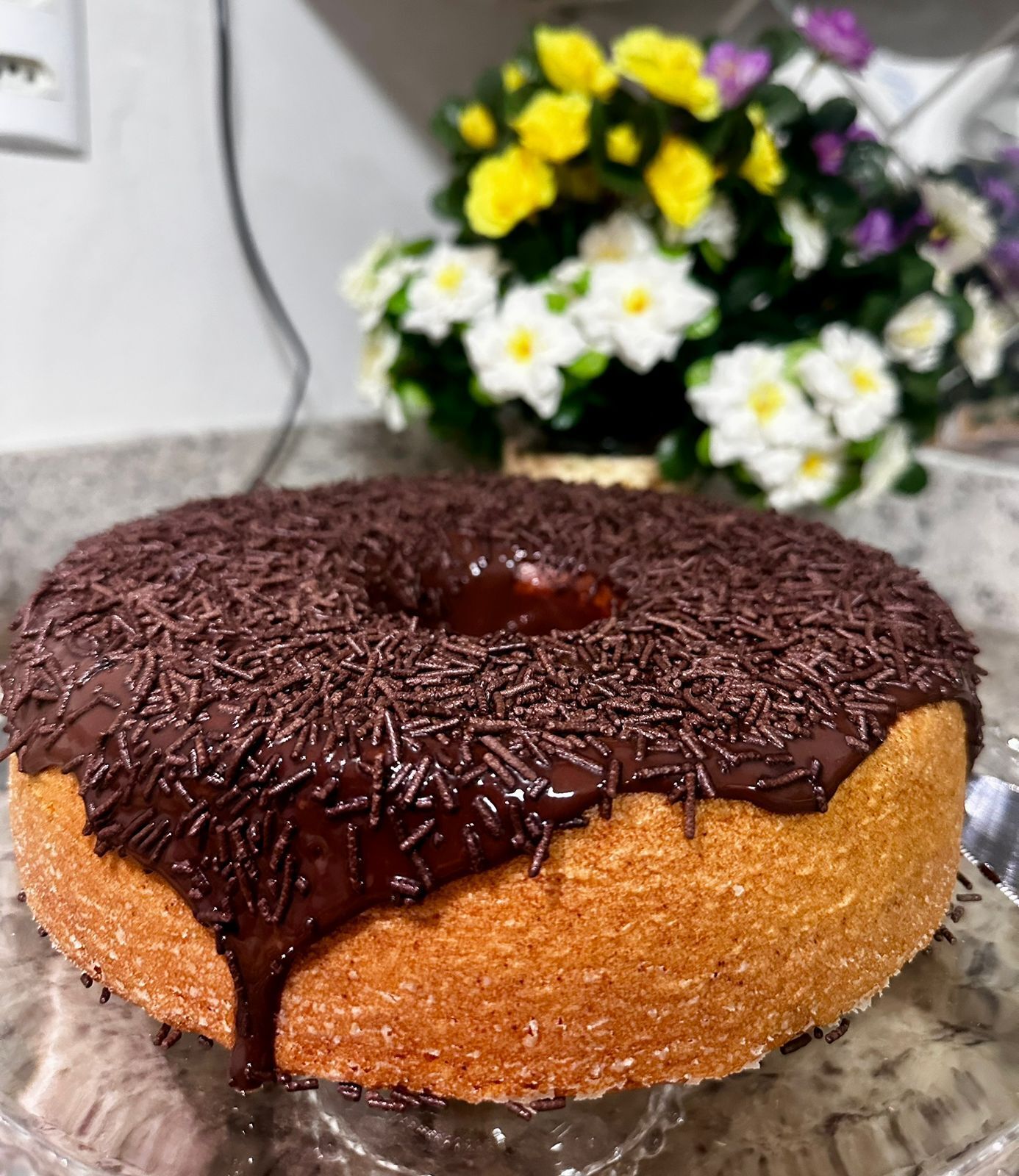 Un pastel bundt bañado en chocolate con grageas sobre un plato de vidrio transparente.