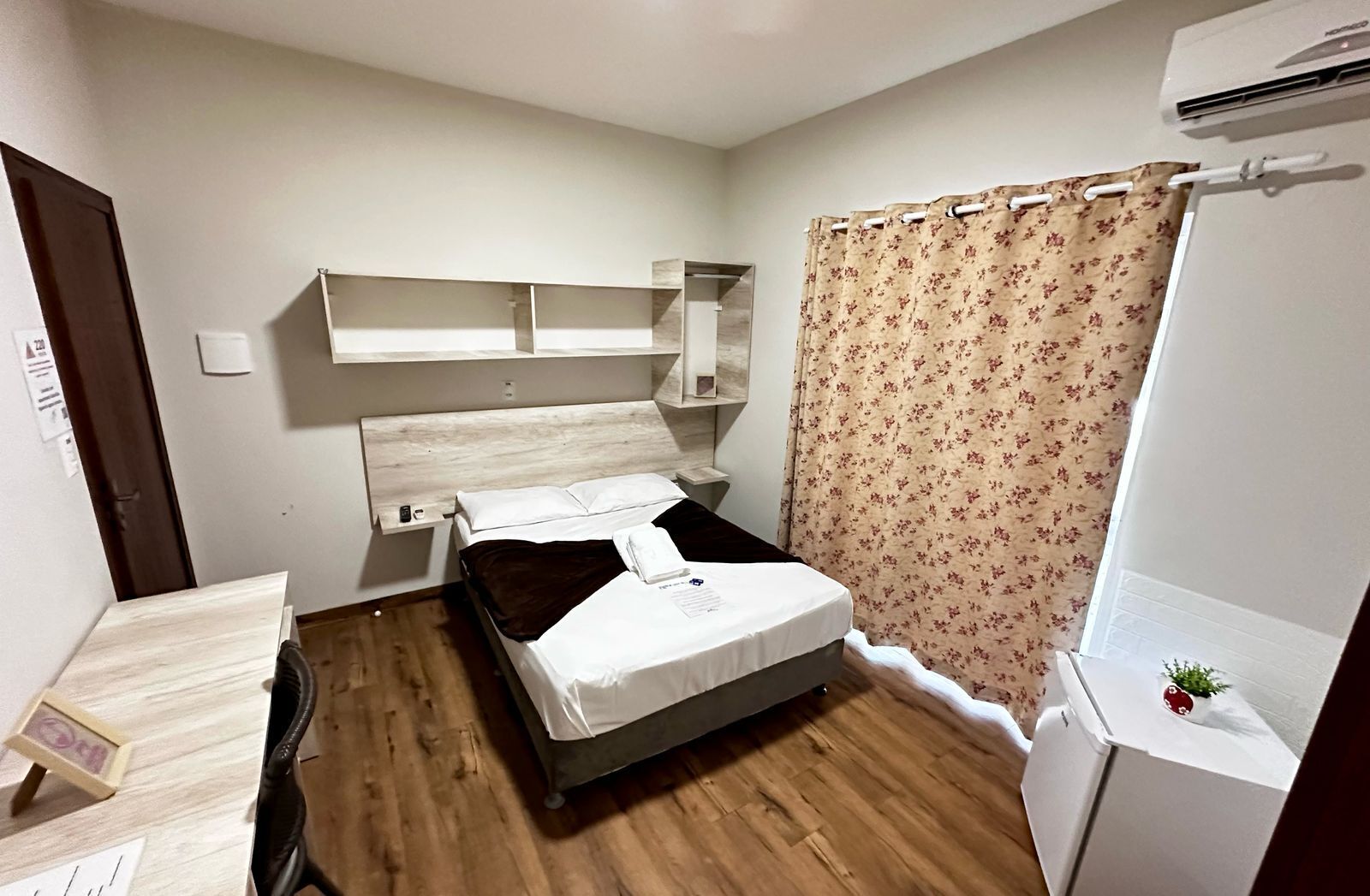 A bedroom with a bed, wooden floor, a small desk, a mirror, and a shower area.