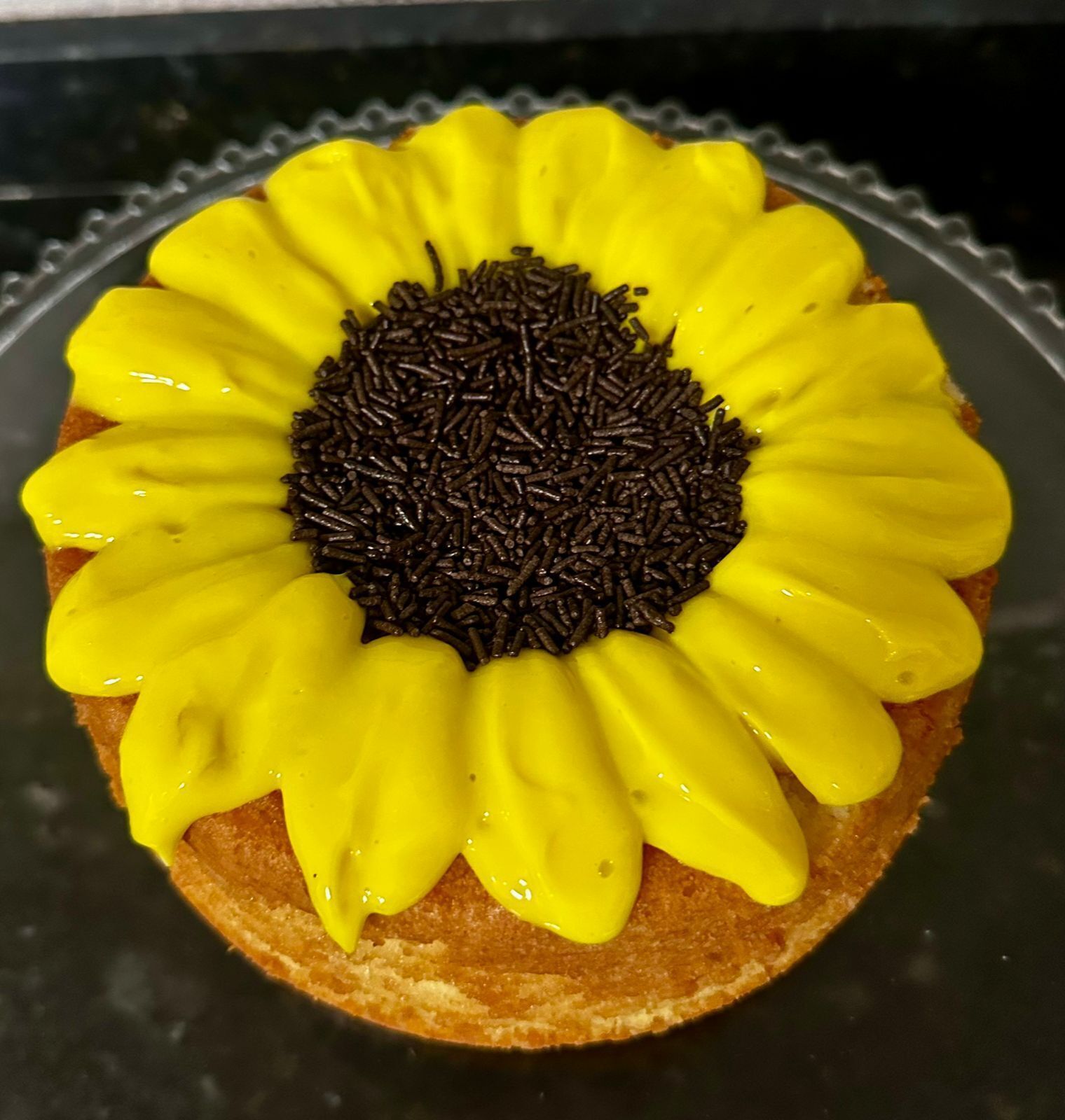 Un pastel redondo cubierto de glaseado amarillo y chispas de chocolate dispuestas para imitar un girasol.