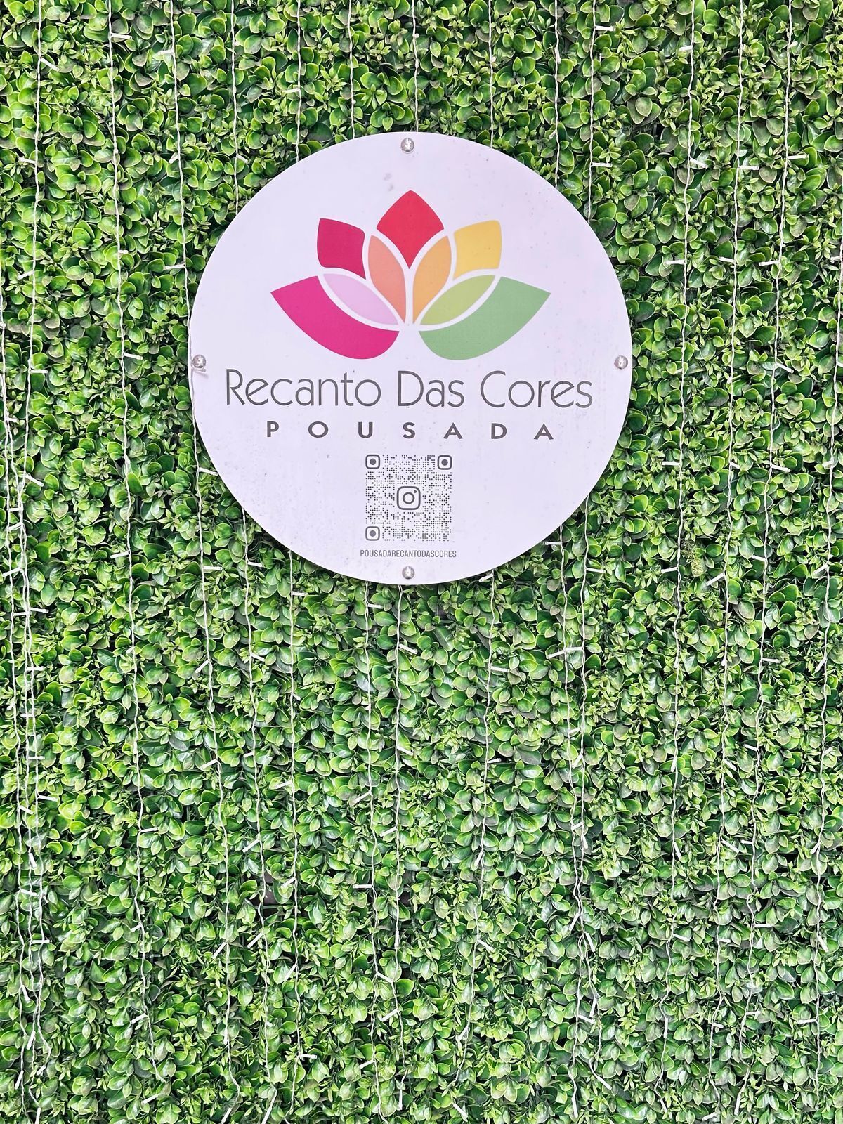 Un cartel con el texto 'Recanto Das Cores' y 'POUSADA' sobre un fondo blanco, rodeado por una pared de follaje verde.