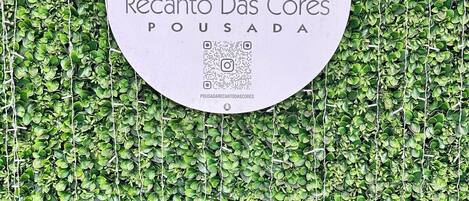 A sign with the text "Recanto Das Cores" and "POUSADA" on a white background, surrounded by a green leafy wall.