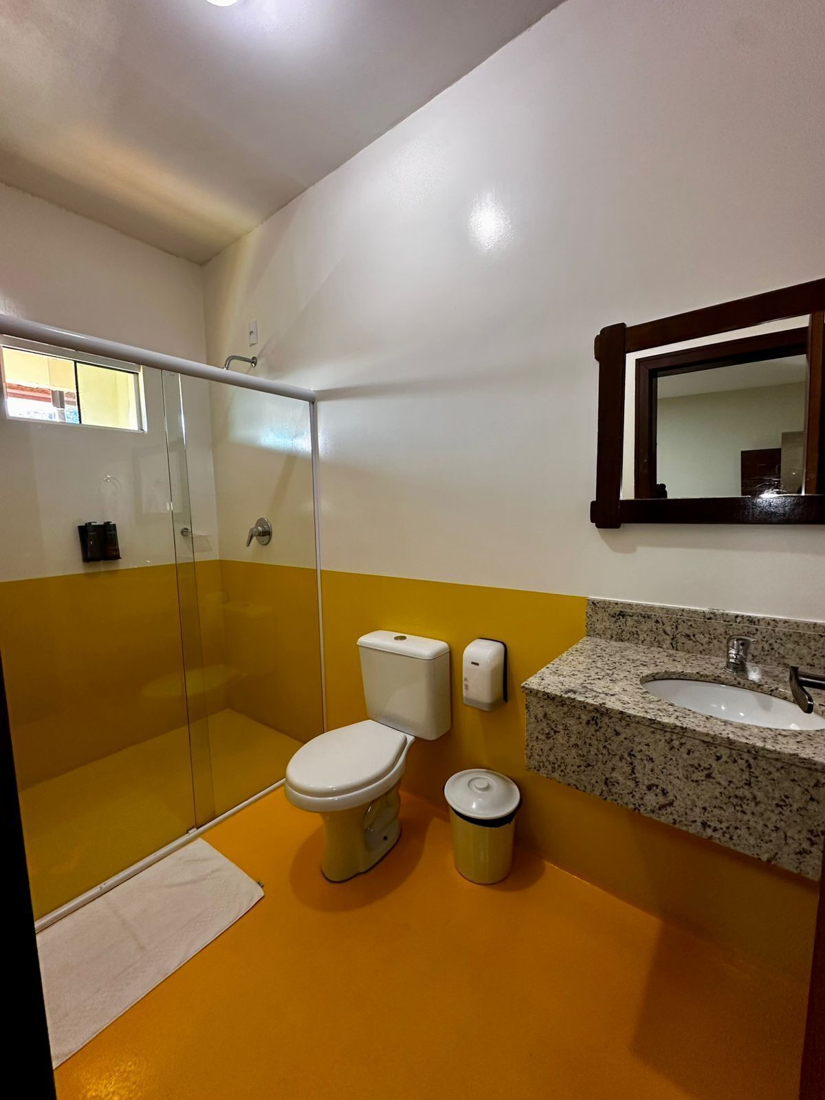 A bathroom with a glass shower enclosure, a toilet, a sink with a granite countertop, and a mirror with a wooden frame.