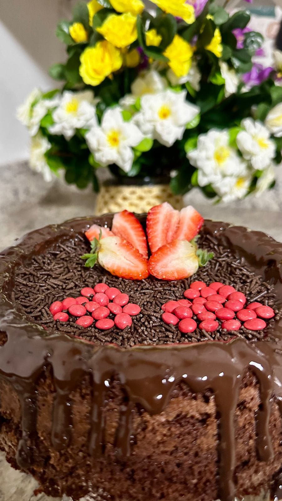 En chokladkaka med jordgubbar och röda godisbitar ovanpå, placerad på ett bord med en blomsterbukett i bakgrunden.